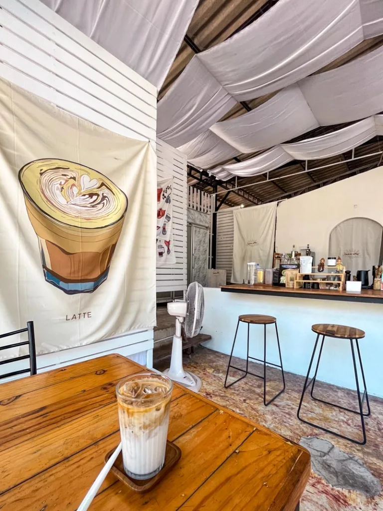 an iced latte on a wooden table with a view of a white coffee bar with wooden stools at sunrise coffee shop in koh tao, thailand