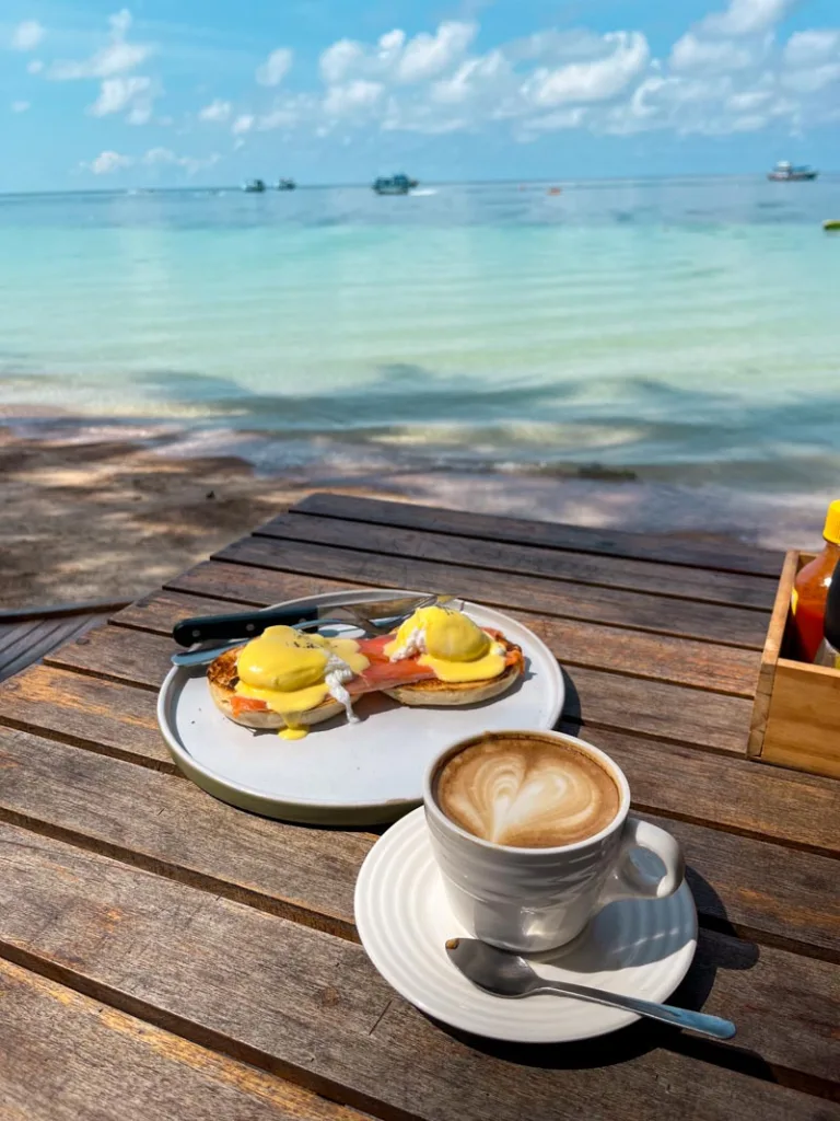a plate of salmon eggs benedict and a latte on a wooden table by the ocean at cafe culture in koh tao, thailand