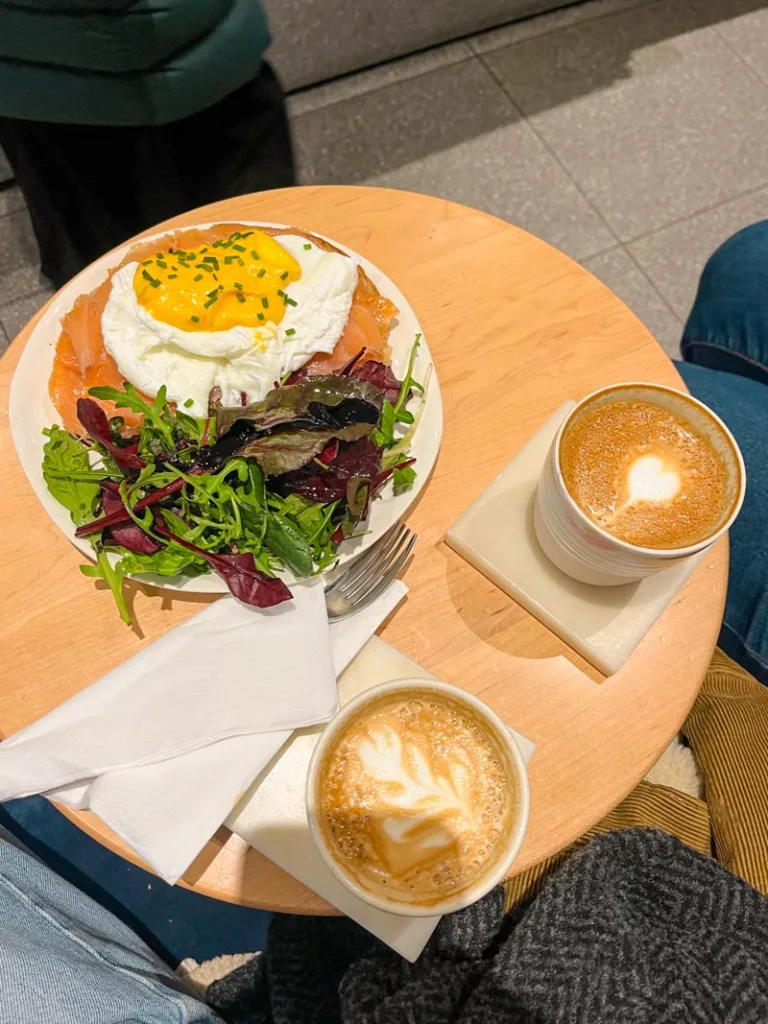 poached egg on a salmon toast and a side salad on a plate, two capuccinnos, at guillam coffee shop in london