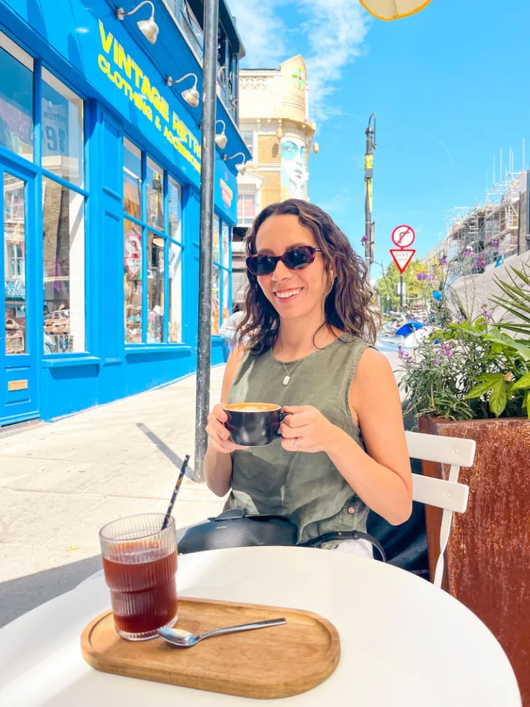 woman smiling holding a cup of coffee at an outdoor table at lancaster brew in notting hill, london