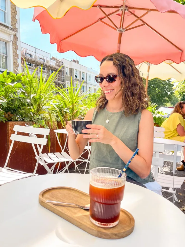 woman holding a latte, sitting on an outdoor table at lancaster brew coffee shop in notting hill, london