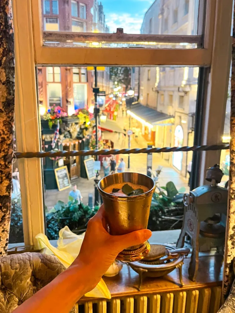 hand holding a cocktail in a metal glass with a view of covent garden out the window at mr foggs gin parlour in london